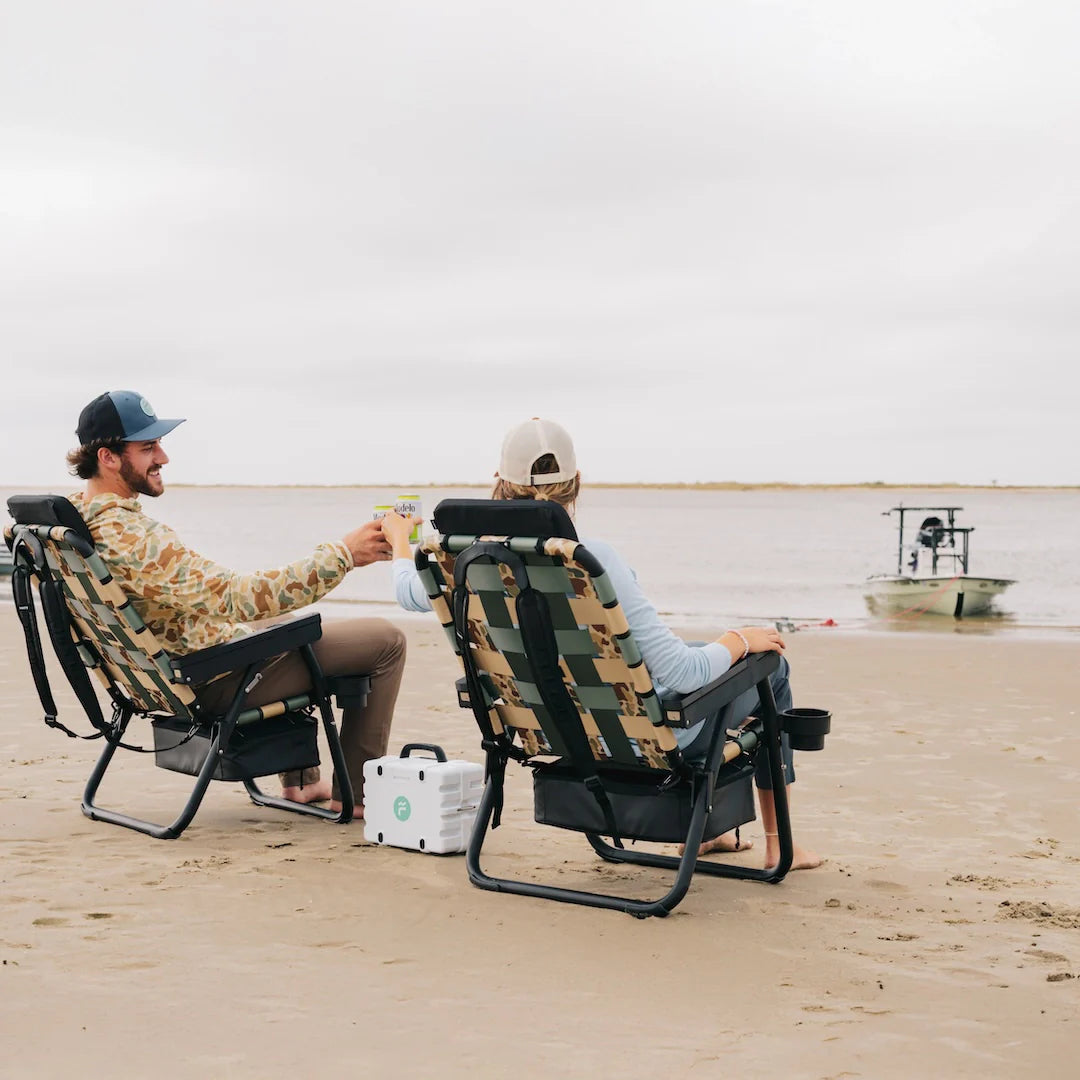 Two friends relaxing on the beach in PARKIT Free Fly Eclipse Recliners in Vintage Camo, with a Turtlebox speaker playing between them and a skiff anchored in the background. These premium folding outdoor chairs feature a built-in cooler, backpack straps, and recline function perfect for beach days, boat trips, or backyard hangs for dads, moms, and outdoor enthusiasts alike.