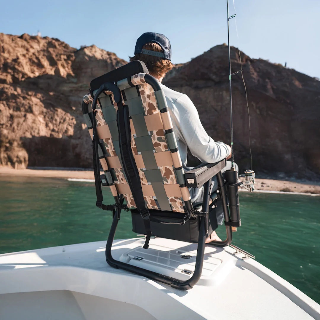 Man fishing from a boat while sitting in the PARKIT Free Fly Eclipse Recliner in Vintage Camo, featuring the Outpost accessory mount holding a fishing rod. This premium folding outdoor recliner combines comfort and function with backpack carry straps, built-in cooler, and marine-grade materials ‚ the perfect fishing chair for dads, outdoor adventurers, and boat lovers seeking durability and all-day relaxation.